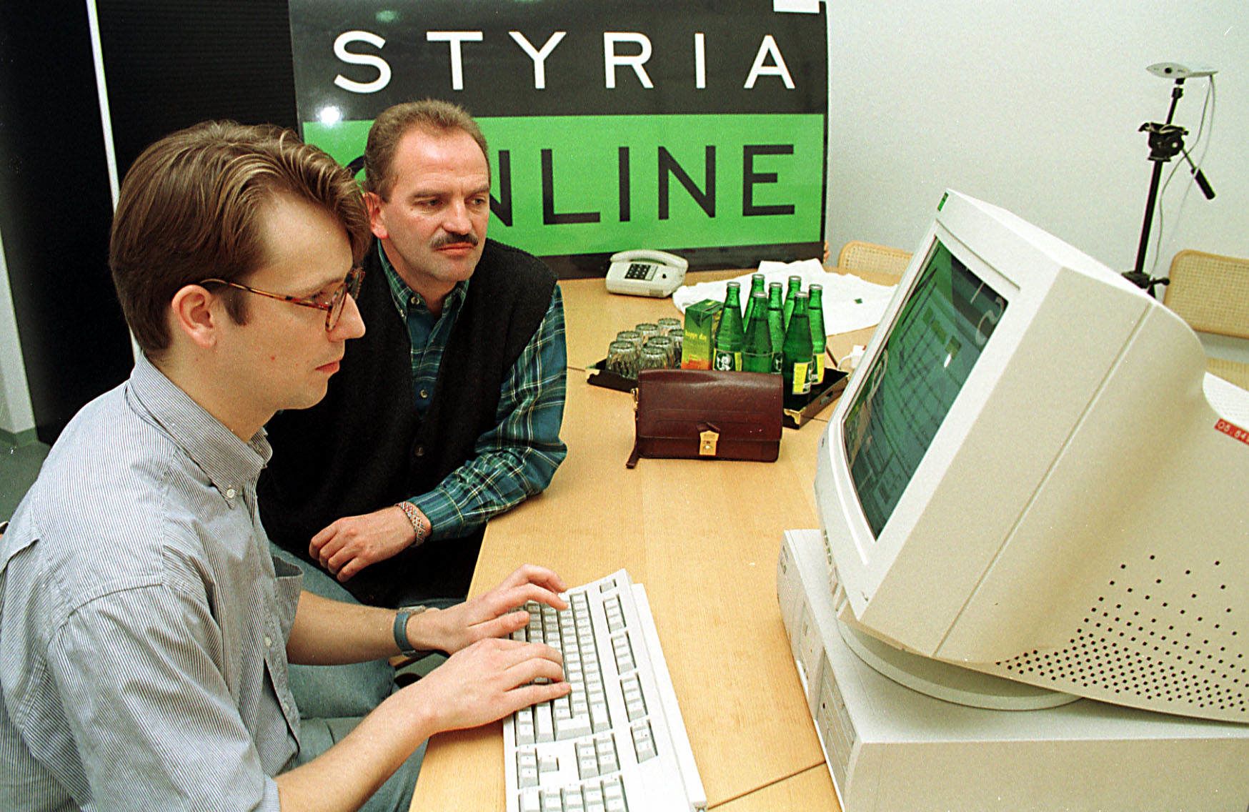 Prohaska und Redakteur Seebacher sitzen vor einem Röhrenmonitor, daneben Getränkeflaschen und eine braune Ledertasche.