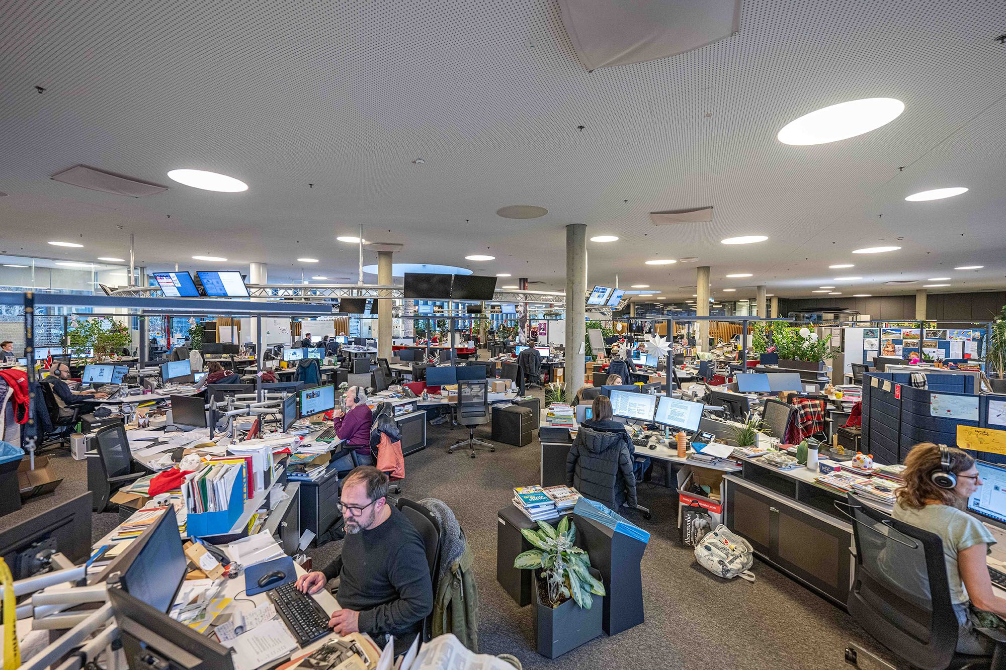 Zu sehen ist ein Großraumbüro mit Dutzenden Arbeitsplätzen. Redakteurinnen und Redakteure sitzen vor Bildschirmen, dazwischen stapeln sich Bücher und Zeitschriften. Auch von der Decke hängen zahlreiche Monitore.