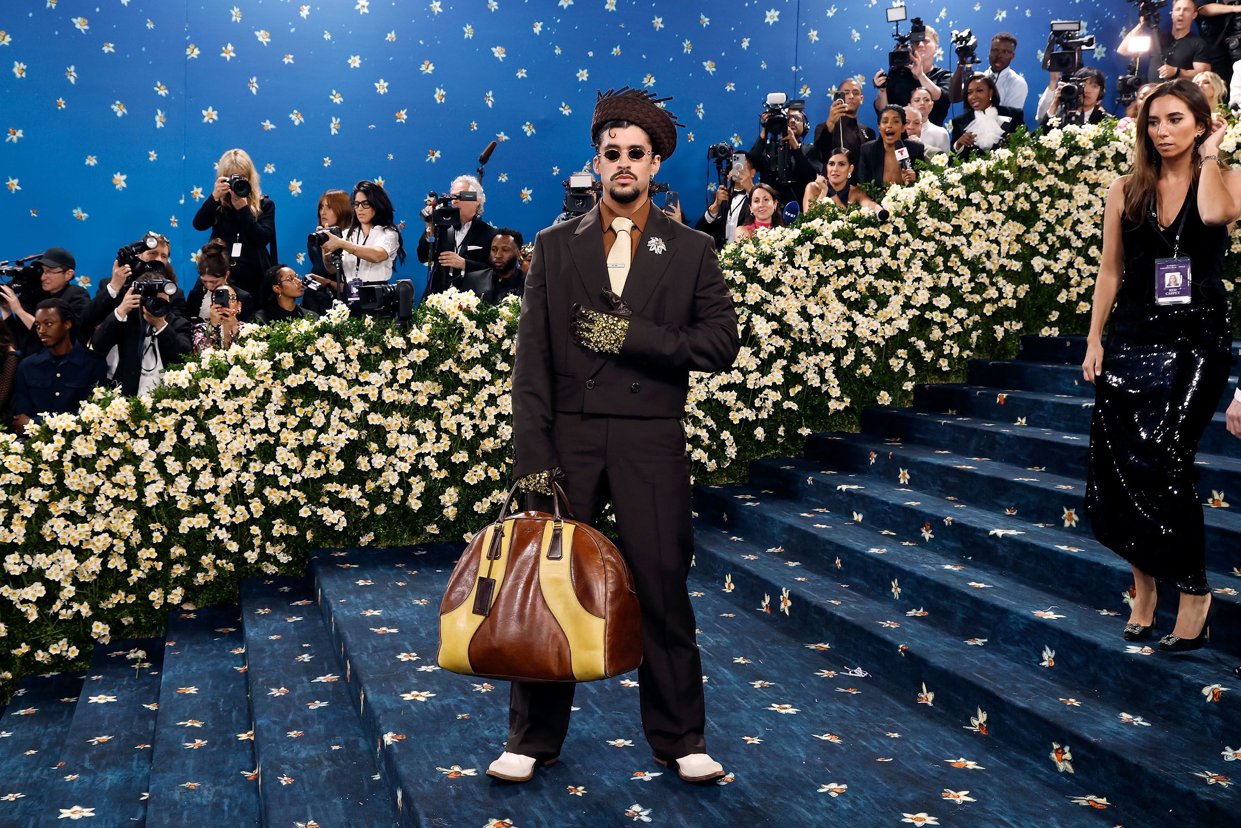 Musiker Bad Bunny wird auf einer Art Red Carpet, der blau ist, fotografiert. Er trägt eine Handtasche, einen Hut und auffällige Handschuhe. Im Hintergrund sind Blumen zu sehen.